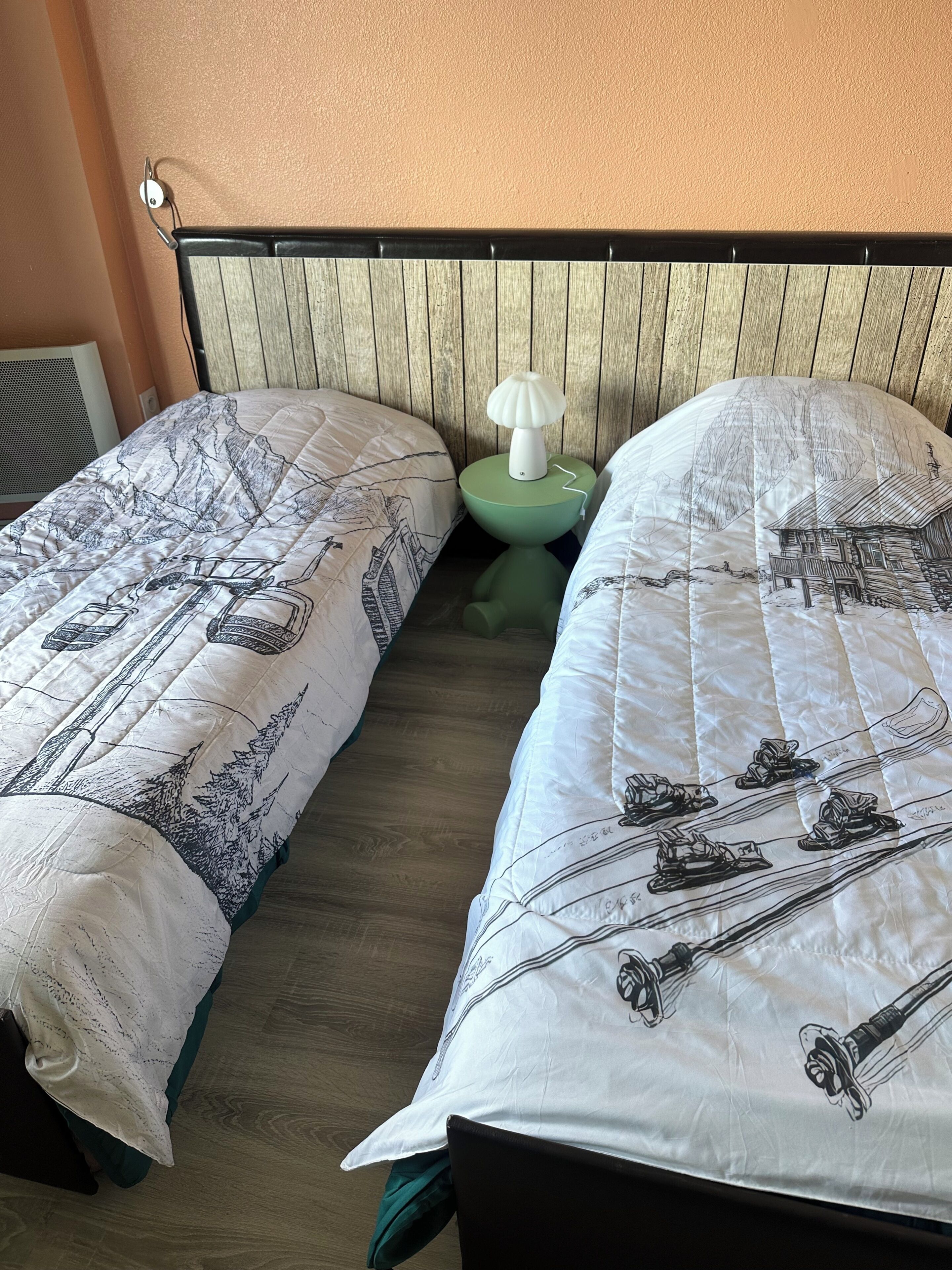 A bedroom with two beds, each with a white duvet featuring black illustrations, a green nightstand with a white lamp, and a wooden headboard.