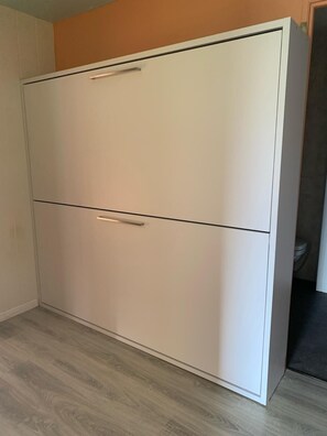 Ein weißer Kleiderschrank mit zwei Türen und Griff in einem Zimmer mit hellem Holzboden und einem teilweisen Blick auf eine Toilette.