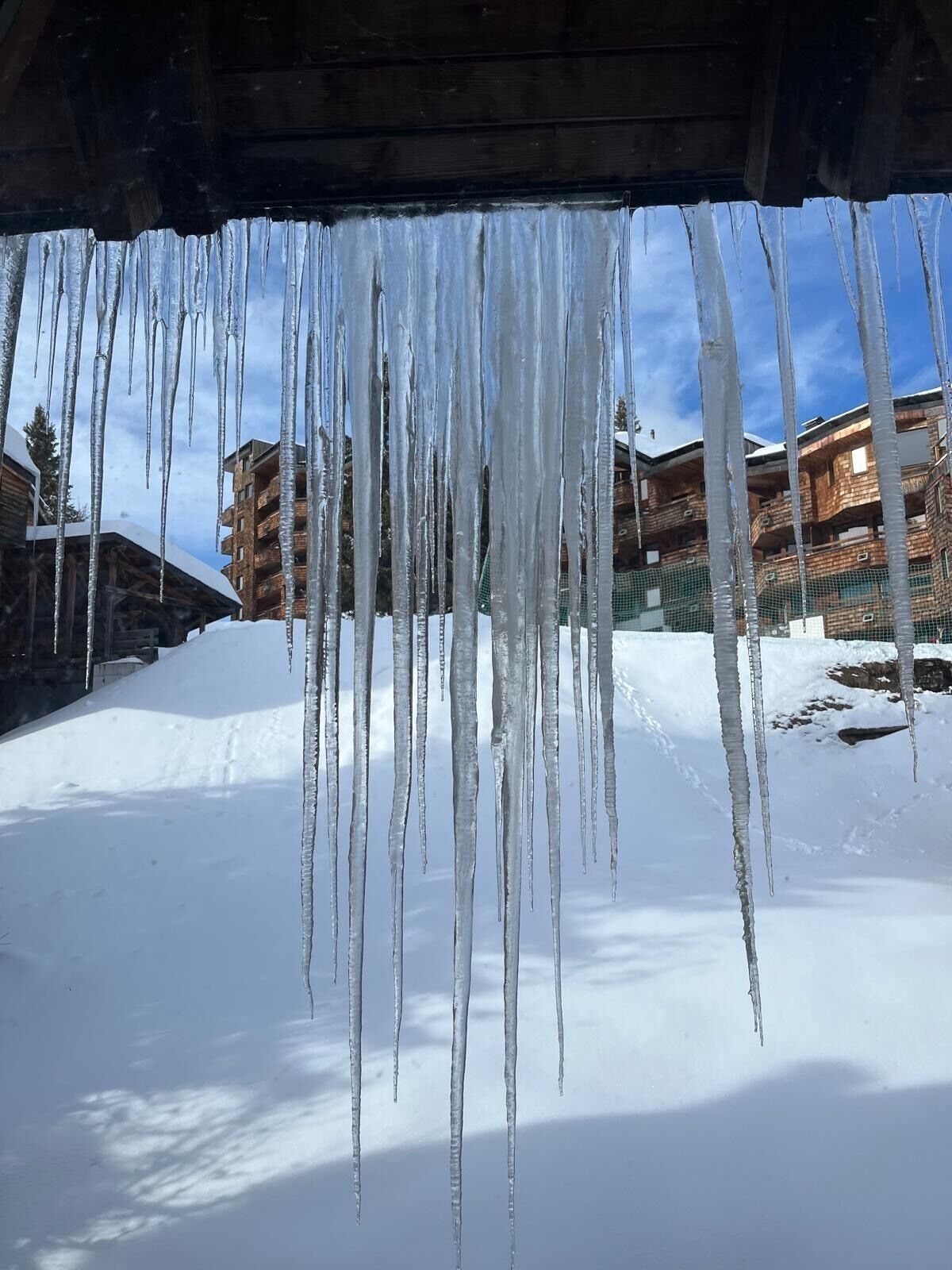 Des glaçons pendent d’une structure en bois, le sol est recouvert de neige et on aperçoit des bâtiments au loin.
