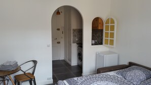 Een kamer met een bed, een tafel met stoelen, een wasmachine en een keuken, zichtbaar door een boog.