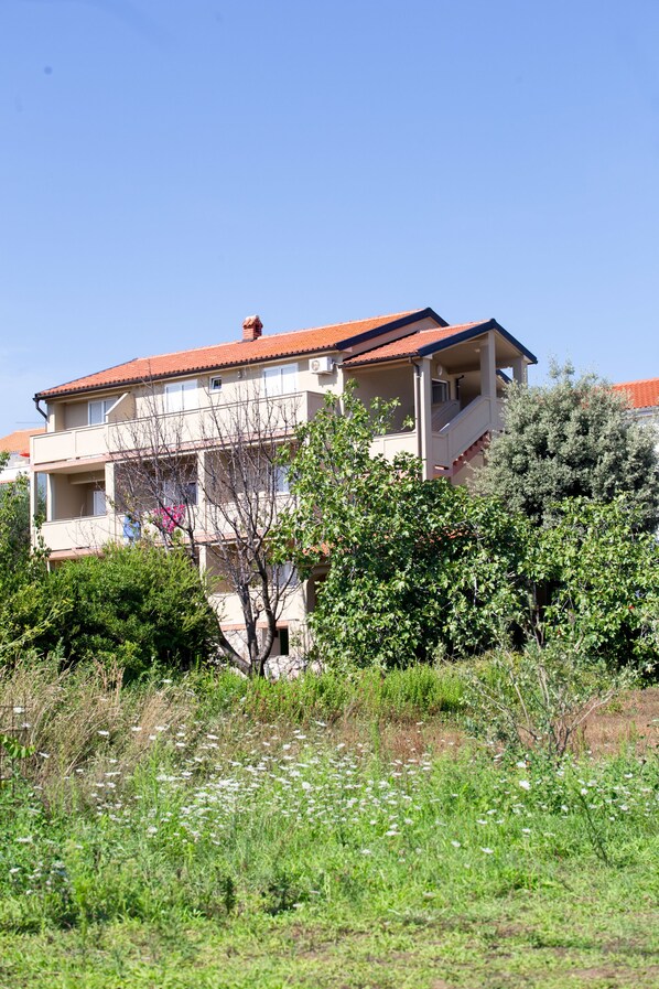 A multi-story building with balconies, surrounded by greenery and a clear sky.