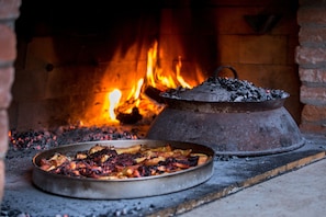 A traditional brick oven with a fire burning inside, a metal pan with food on a table in front of it, and a large pot on the oven's side.