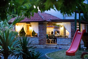 Een gebouw van steen met een dak van rode tegels, een terras met stoelen en een tafel, en een rode glijbaan.