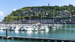 Uma marina com vários barcos atracados, um cais em primeiro plano e uma colina com edifícios e uma torre ao fundo.