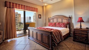 A bedroom with a large bed, a ceiling fan, a nightstand with a lamp, and a view of the city through a balcony. - Bella Sirena 1BR 303 by Casago (Puerto Peñasco)