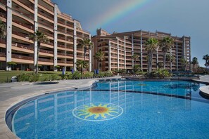 Ein Hotel mit großem Pool, der ein Sonnendesign zeigt, umgeben von Palmen und blauem Himmel.