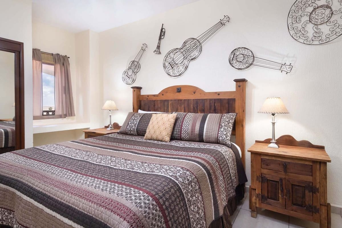 Ein Schlafzimmer mit einem hölzernen Bett, gemusterten Betttextilien, zwei Nachttischen mit Lampen und an der Wand befestigten Gitarren.