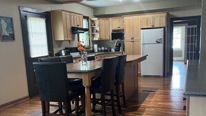 Una cocina con armarios de madera, una isla central con sillas, un refrigerador y un ventanal con persianas.