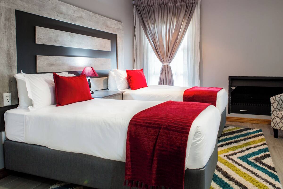 A modern hotel room with a large bed, two red pillows, a grey headboard, a patterned armchair, and a fireplace.