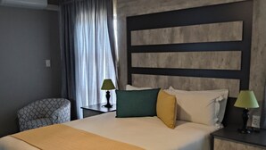 Une chambre d’hôtel moderne avec un grand lit, une table de chevet avec une lampe, un fauteuil à motifs et une fenêtre avec des rideaux.