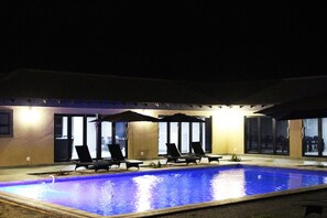 An outdoor swimming pool with lounge chairs and umbrellas, surrounded by a building with large windows. - Sagwadi Hotel (Bushbuckridge)