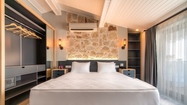 A modern hotel room with a large bed, built-in wardrobe, stone accent wall, and a large window with curtains. - Isola12 (Kas)