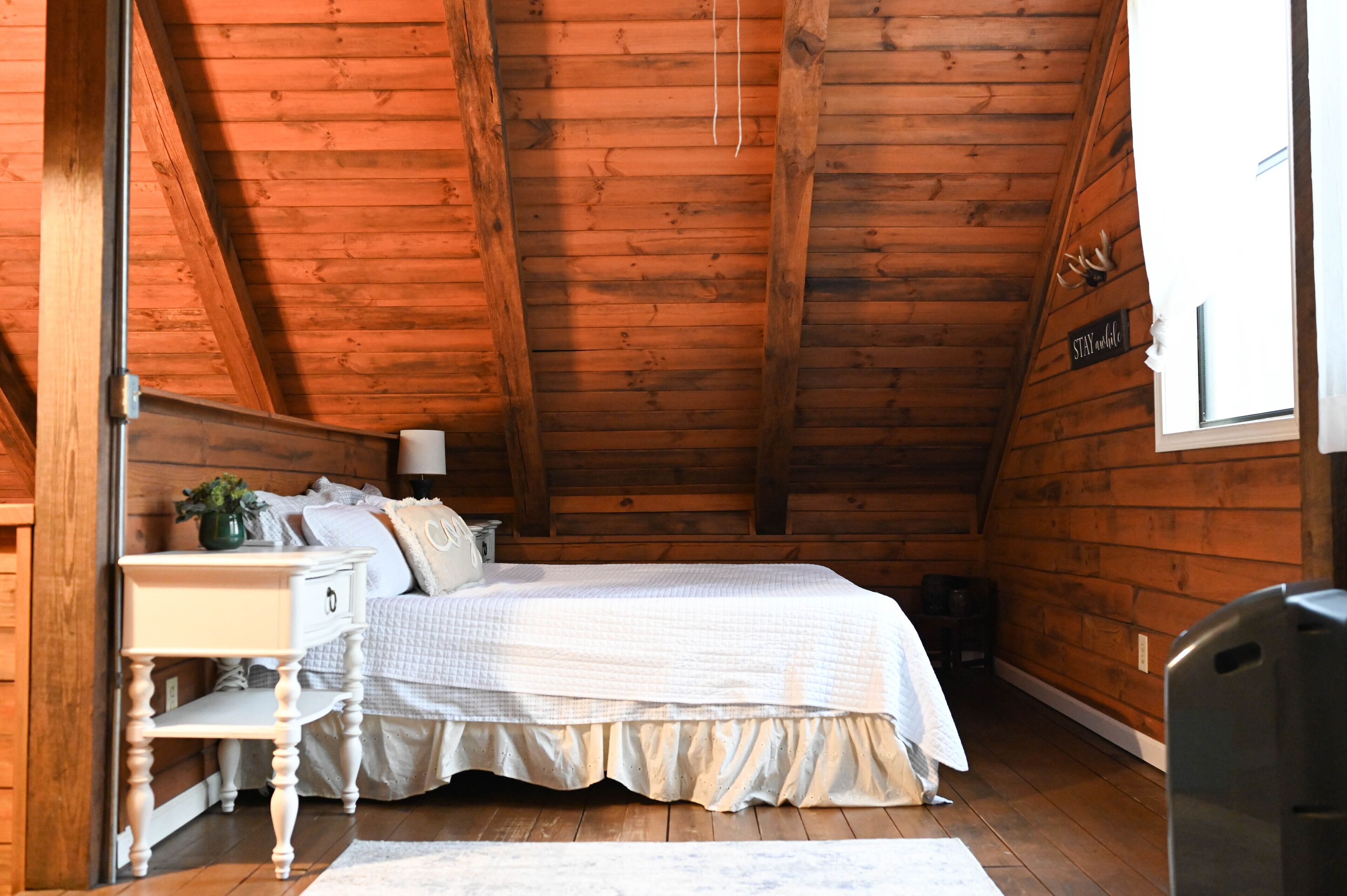 A cozy bedroom with a wooden ceiling, a bed with a white bedspread, a nightstand with a lamp, and a window with a sign saying "STAY warm".