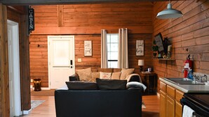 Ein gemütliches Blockhäuschen-Interieur mit einer Couch, Holzwänden, einer Tür, einem Fenster mit Vorhängen, einem kleinen Tisch mit Lampe und einer Küchenzeile mit Spüle und Schränken.