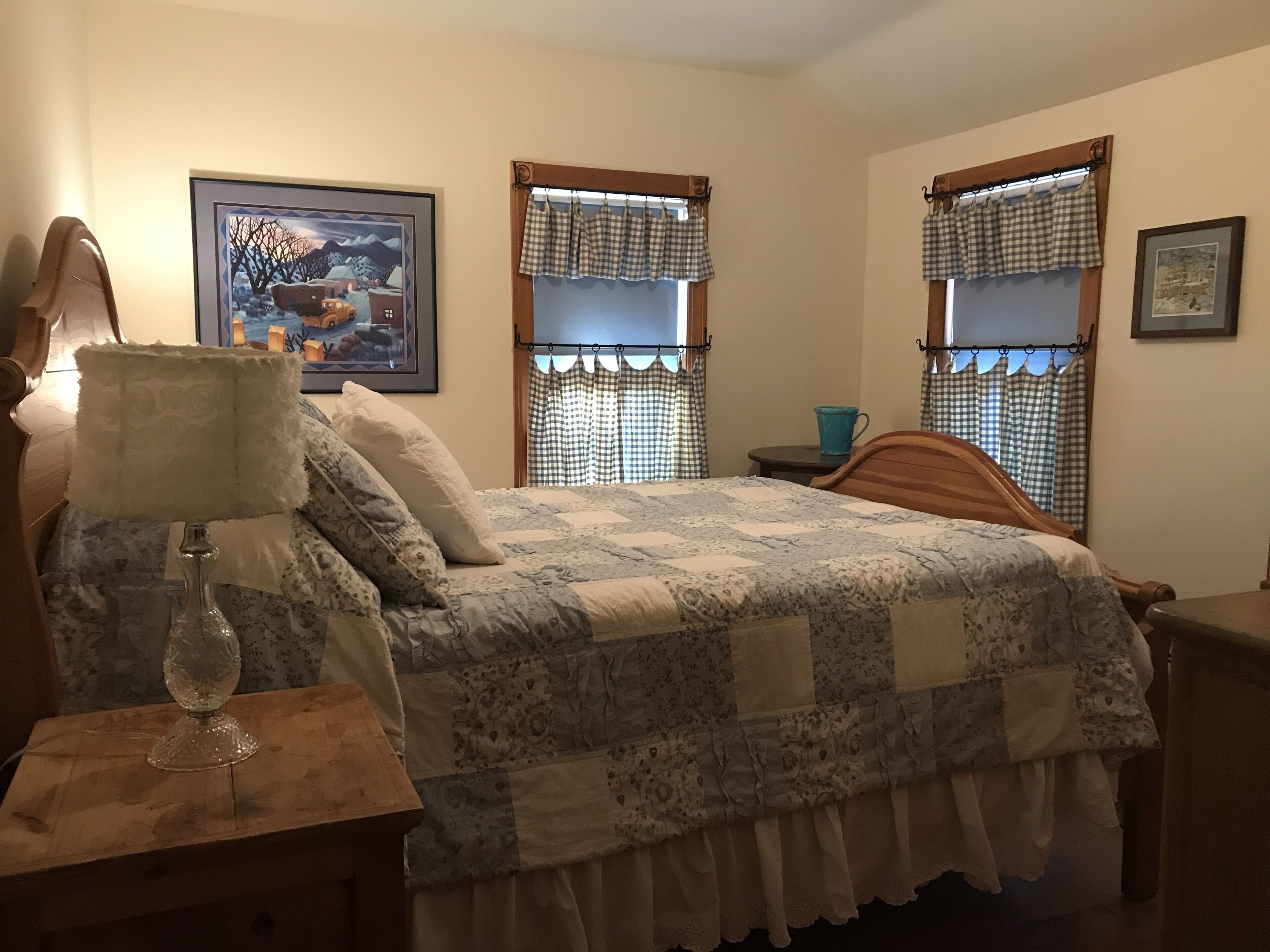 A bedroom with a wooden bed, a bedside table with a lamp, a framed picture on the wall, and checkered curtains on the windows.