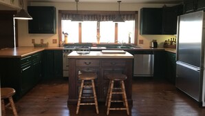 A kitchen with dark wooden cabinets, a stainless steel refrigerator, a wooden island with stools, and a window with curtains. - Beautiful 4br home, village of Bolton Landing. Great location! Walk everywhere! (Bolton Landing)