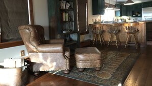 A living room with a leather armchair, ottoman, and a bookshelf. There is a kitchen area with bar stools and a hanging light fixture. - Beautiful 4br home, village of Bolton Landing. Great location! Walk everywhere! (Bolton Landing)