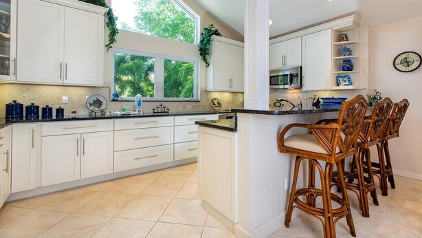 Een keuken met witte kastjes, een centraal eiland met barkrukken, een magnetron en een klok aan de muur.
