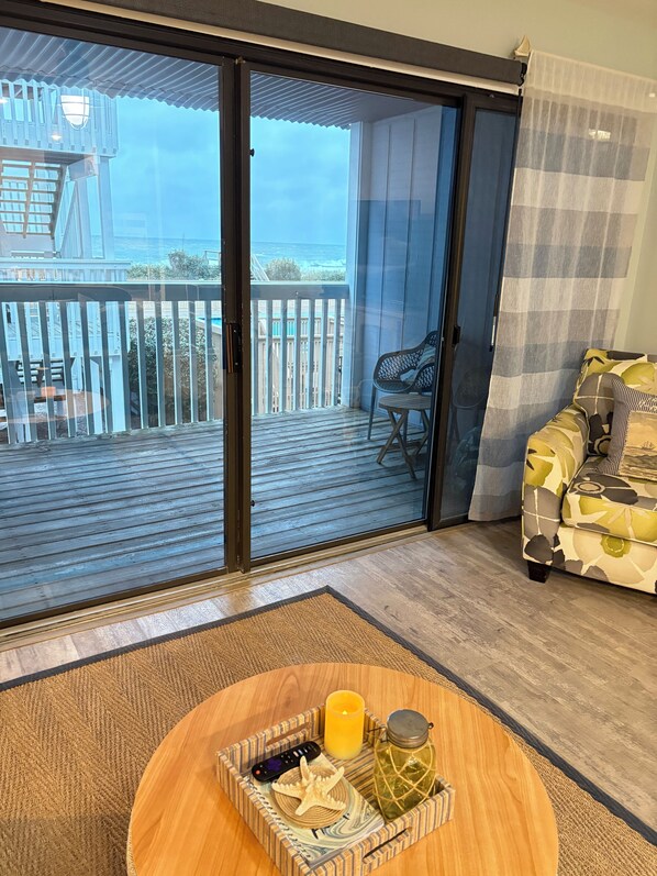 Ett rum med en skjutdörr i glas som leder ut till en balkong med utsikt över havet. Där står ett runt träbord med ett bricka på, och på brickan finns ett ljus, en burk och en fjärrkontroll. En mönstrad soffa står längs väggen.