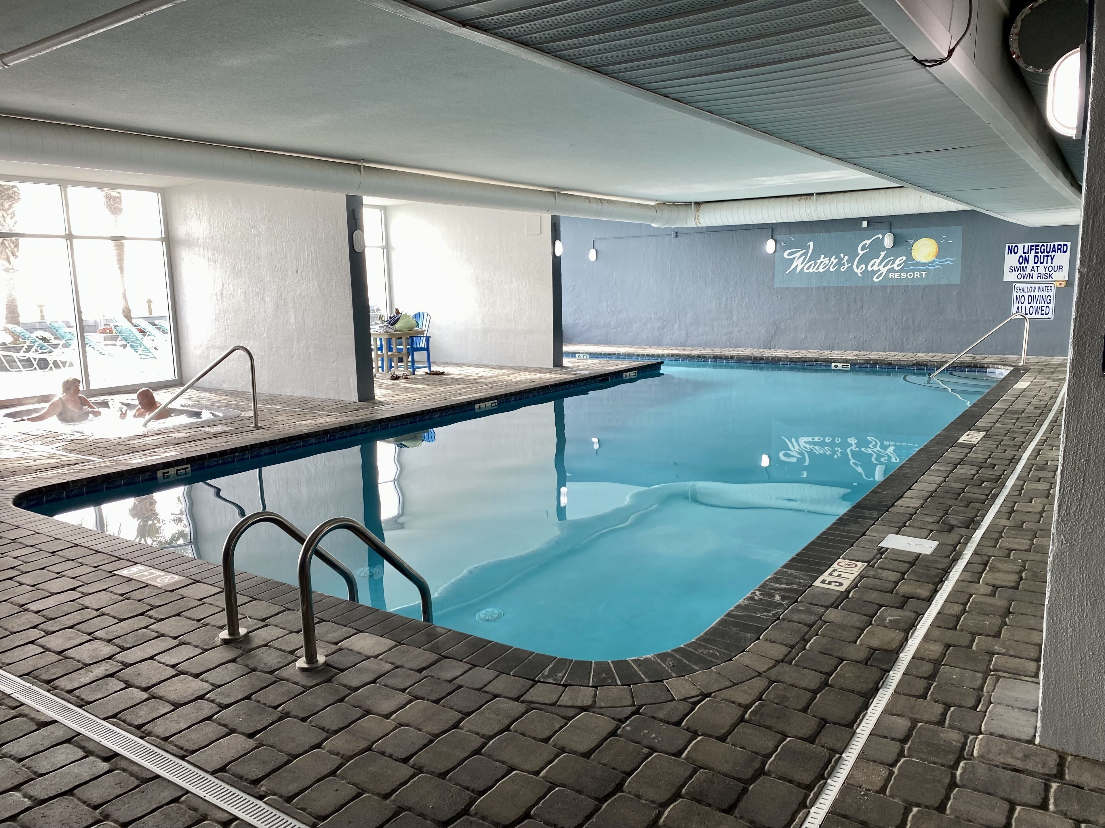 An indoor swimming pool with a tiled deck, a lifeguard sign, and a wall with the text "Water's Edge Resort".