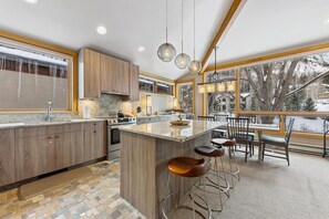 A modern kitchen with a large island, bar stools, and a view of a snowy landscape through large windows. - Creek‑Side Millrace Three‑Bedroom Retreat (Vail)