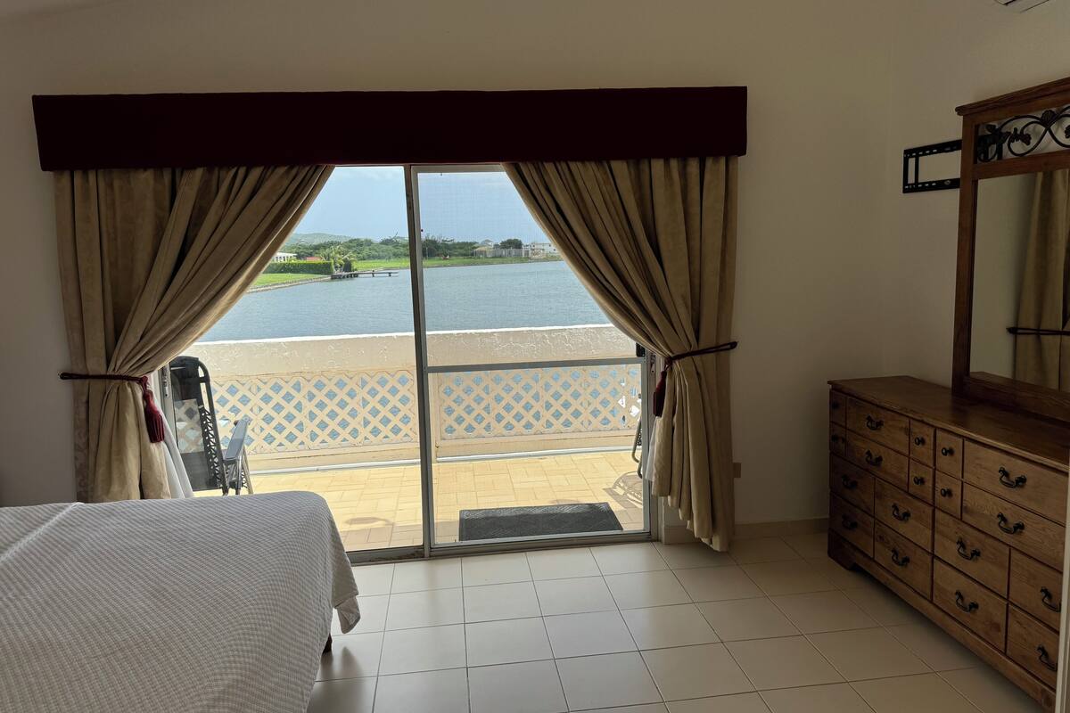 A hotel room with a bed, a wooden dresser, and a view of a body of water through a glass door.