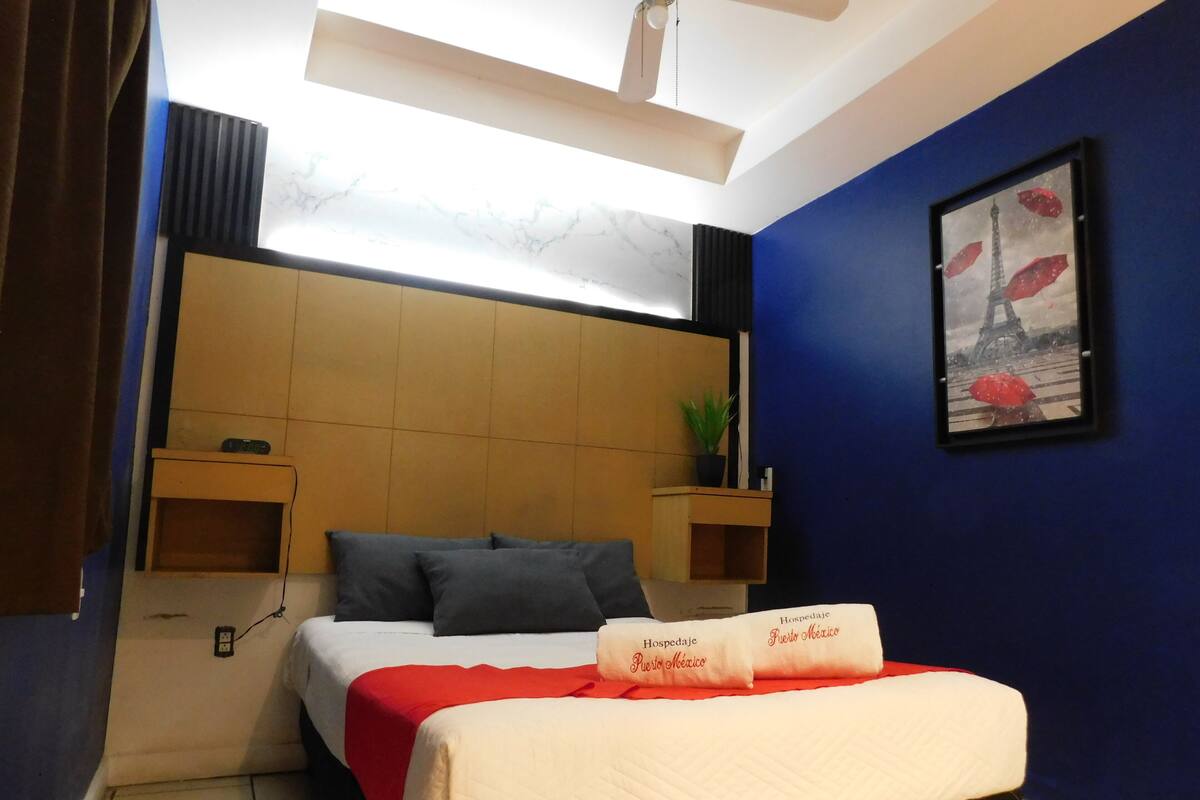 A hotel room with a bed, a headboard, a bedside table, a nightstand, a wall-mounted shelf, a framed picture, and a ceiling fan.