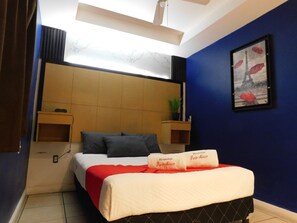 Una camera d'hotel con un letto, una testiera, un comodino, una mensola a muro, un quadro incorniciato e una ventola a soffitto.