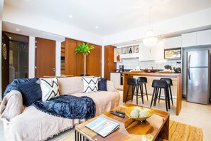 A modern living room with a sofa, a coffee table, and a kitchen area with a refrigerator and bar stools.