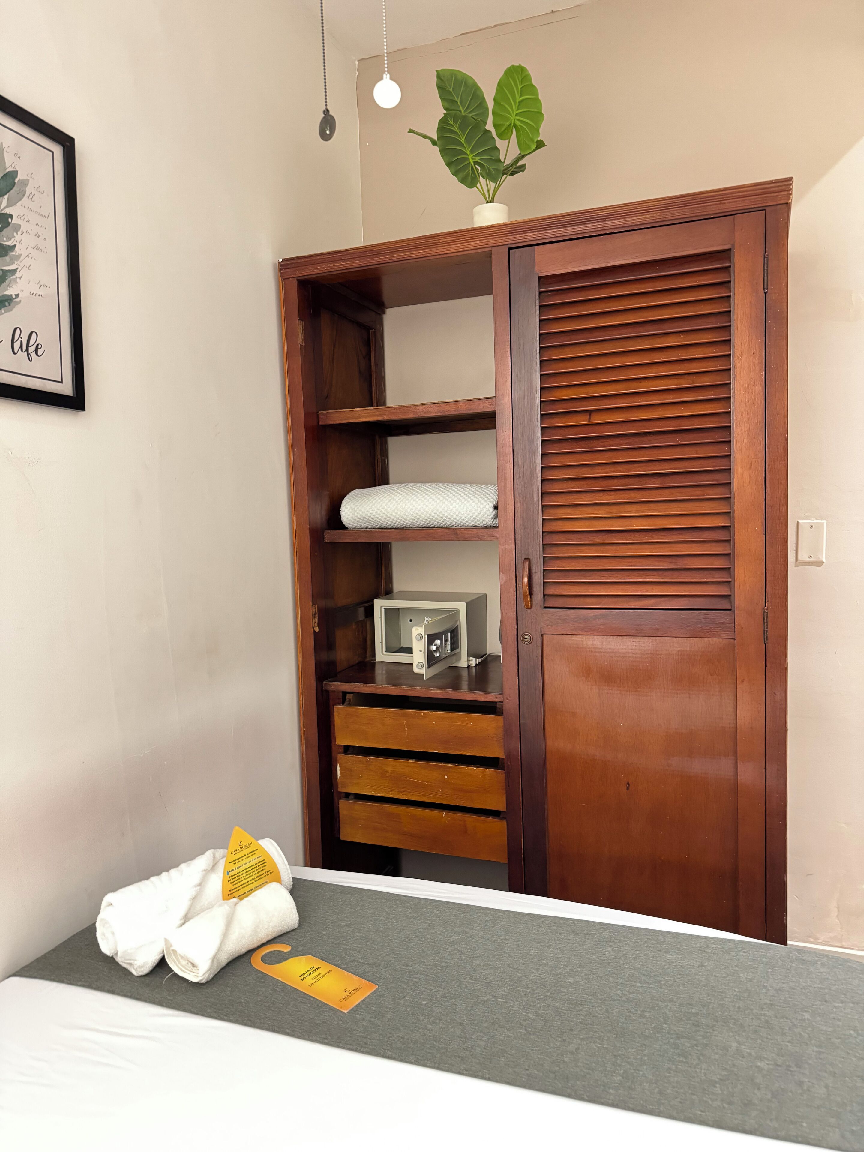 Un dormitorio con un armario de madera, una cama con colchón gris y una mesita de noche con una toalla y una tarjeta de identificación.
