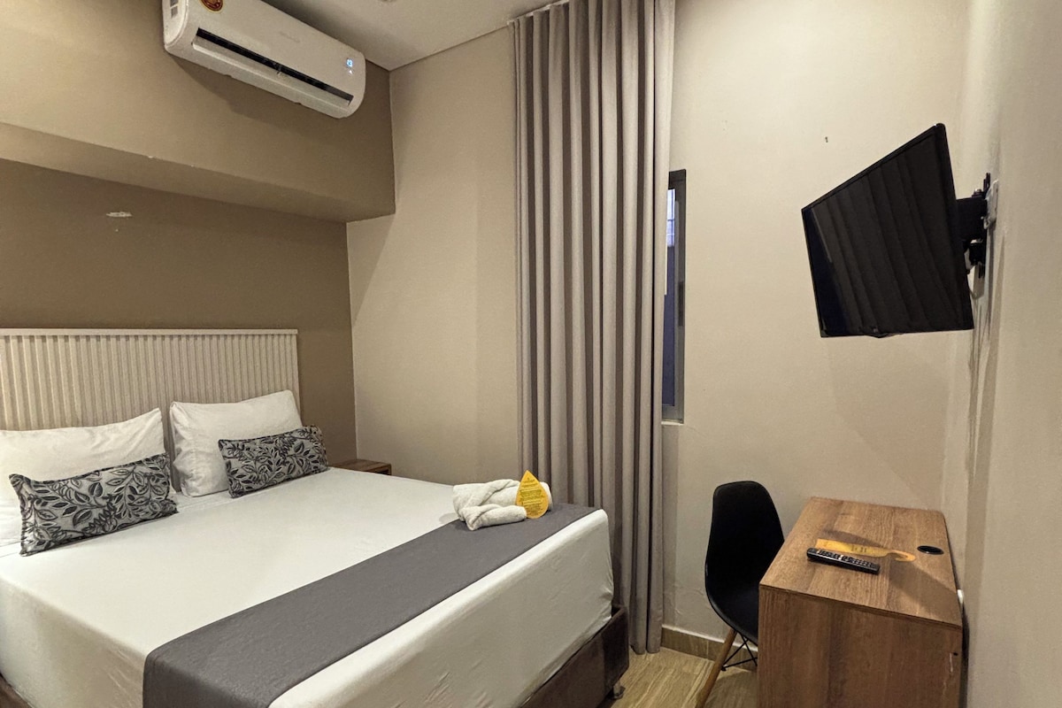 A hotel room with a bed, a desk, a chair, an air conditioning unit, a television, and a window with curtains.