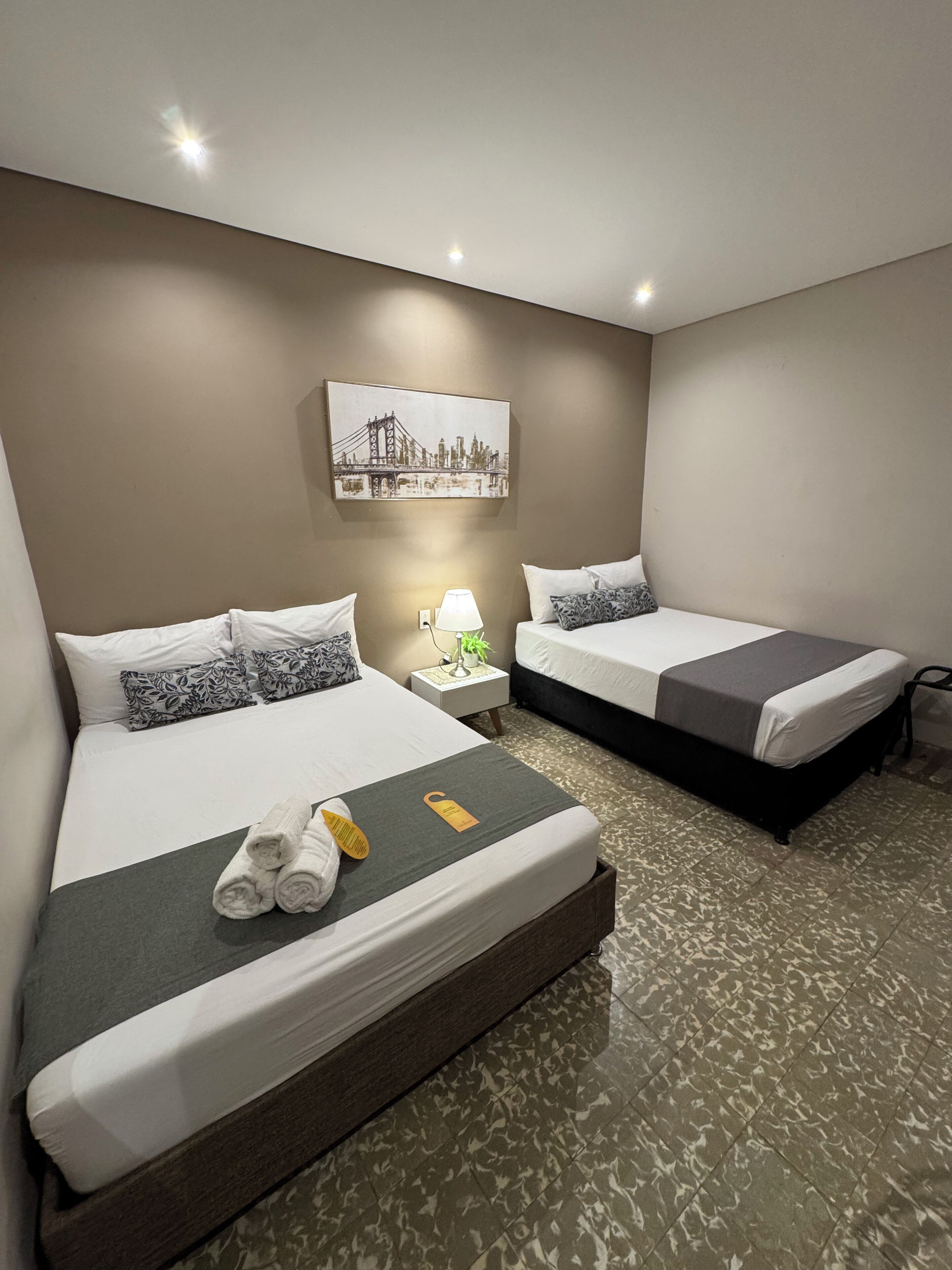 Habitación de hotel con dos camas, una mesita de noche con lámpara, un cuadro en la pared y un par de toallas sobre la cama.