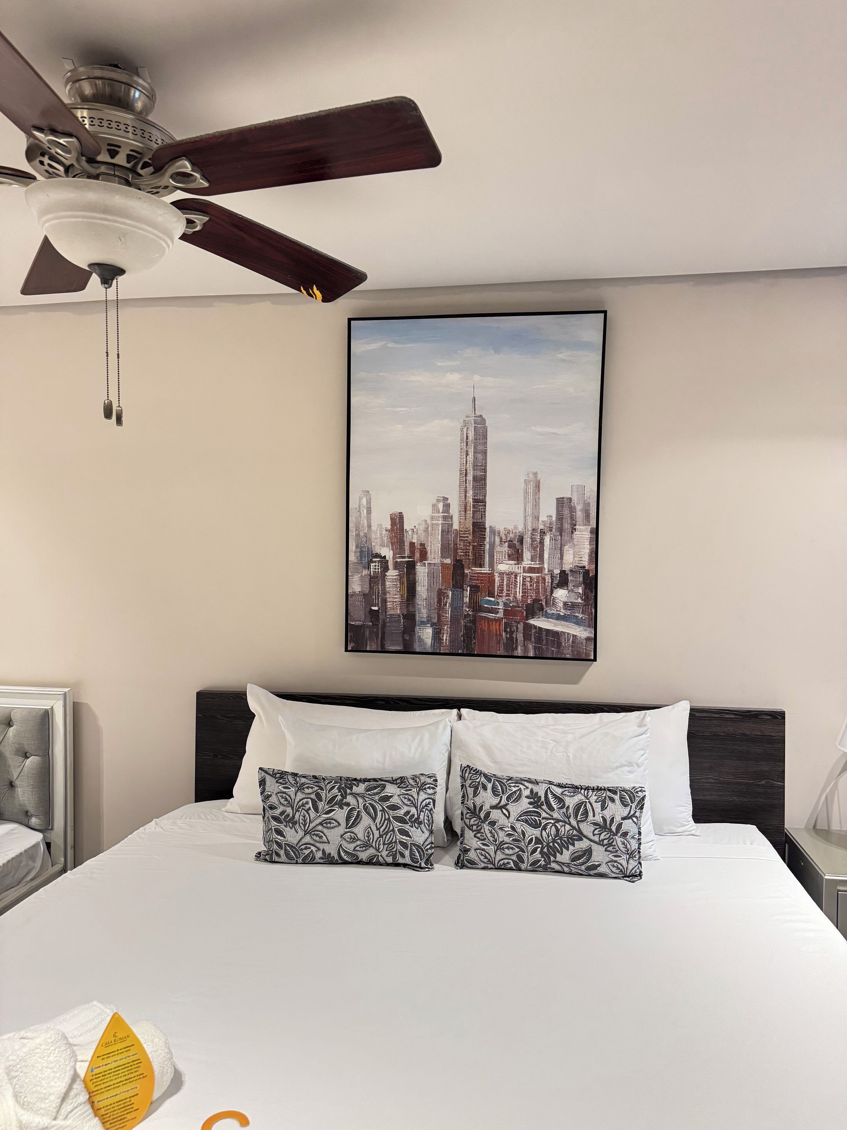 A hotel room with a bed, a ceiling fan, a cityscape painting, and a nightstand with a lamp.