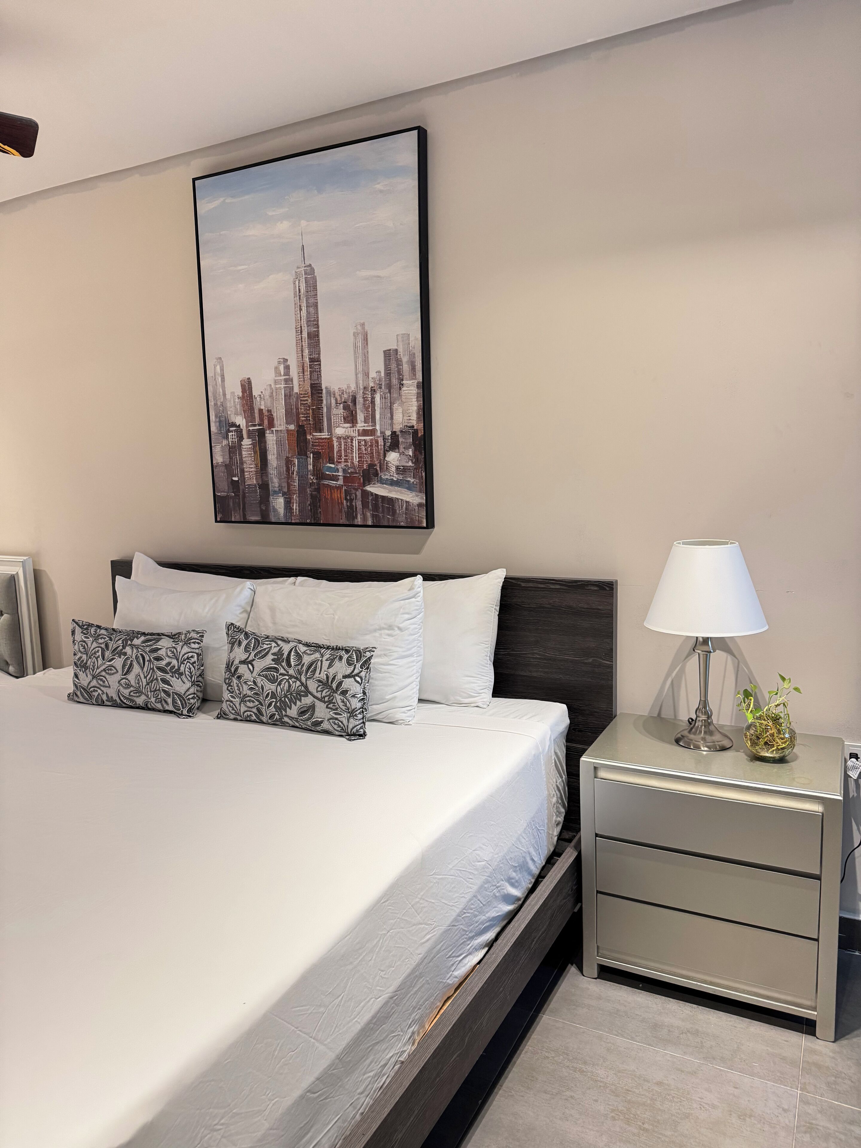 A modern bedroom with a cityscape painting, a bed with white linens and patterned pillows, a bedside table with a lamp and a plant, and a nightstand with drawers.
