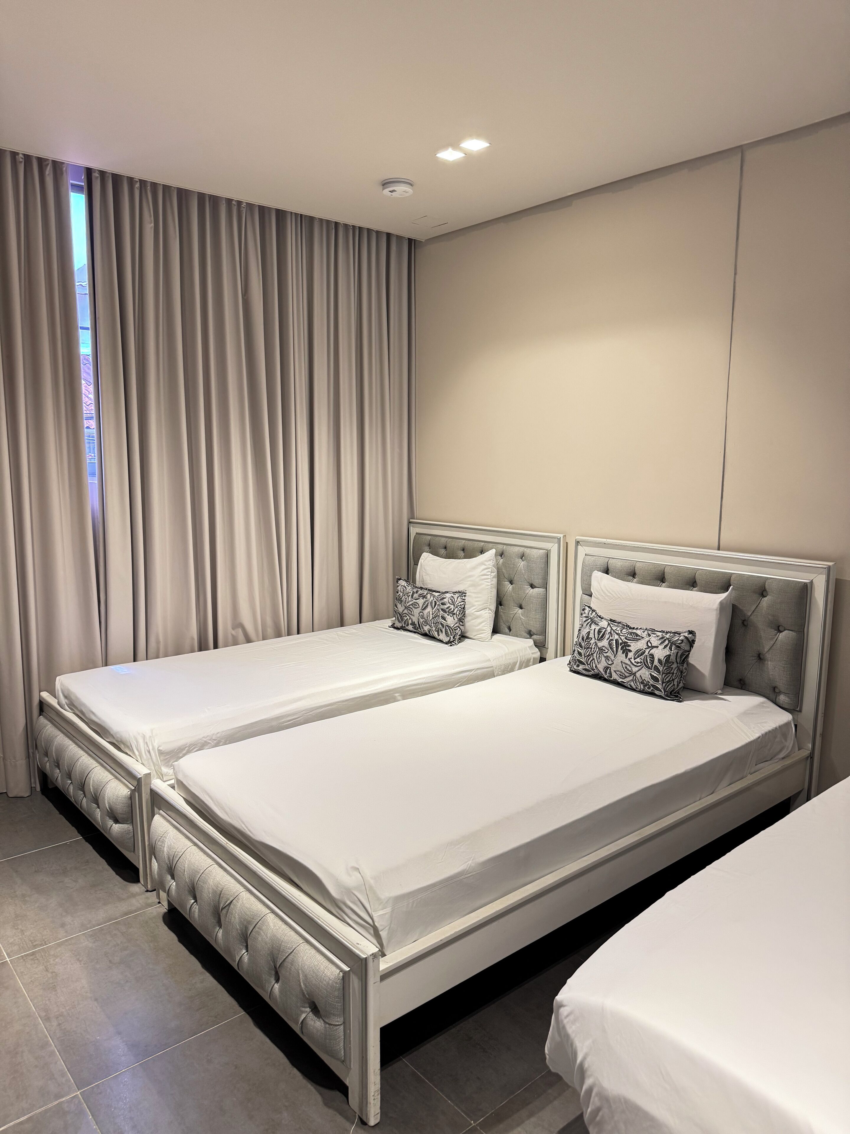 A modern bedroom with two beds, a large window with curtains, and a ceiling light.