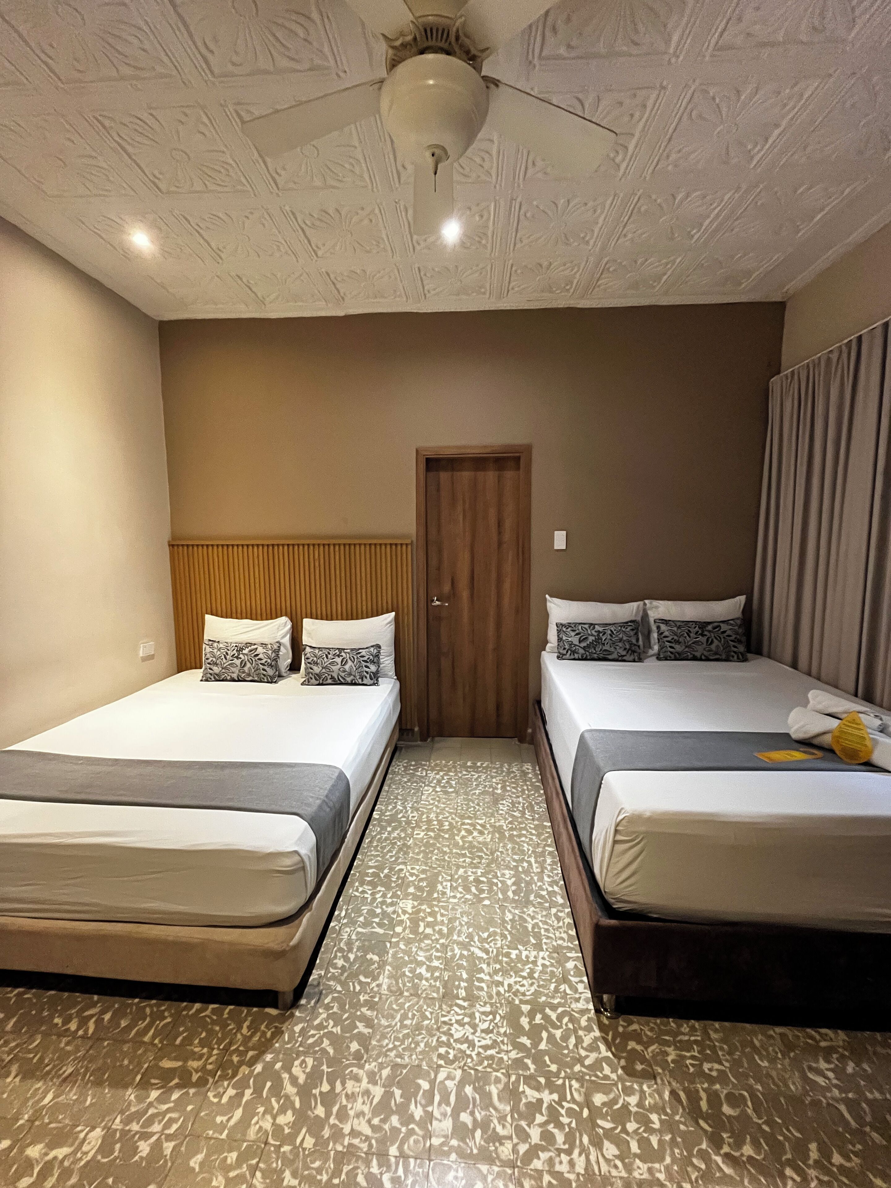 A hotel room with two single beds, a wooden headboard, a patterned floor, and a ceiling fan.