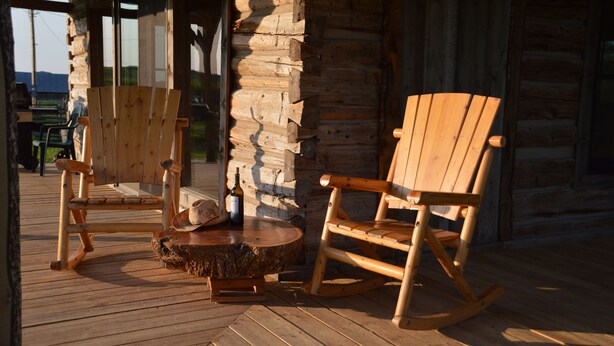 Une chaise à bascule en bois et une chaise en bois sont posées sur une terrasse en bois, à l’extérieur d’une cabane en rondins.