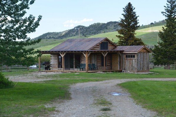 A rustic wooden cabin with a porch and two chairs, surrounded by a grassy area and trees. - Near glacier park. Perfect setting for outdoor enthusiasts. Family friendly. (Dupuyer)