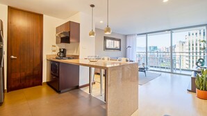 Een moderne keuken met een centraal eiland, inbouwoven en koffiezetapparaat. Er is een zithoek met tafel en stoelen, en een groot raam met uitzicht op de stad.