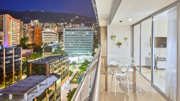 Een balkon met uitzicht op de stad 's nachts, met moderne gebouwen, een eethoek met stoelen en een glazen deur.