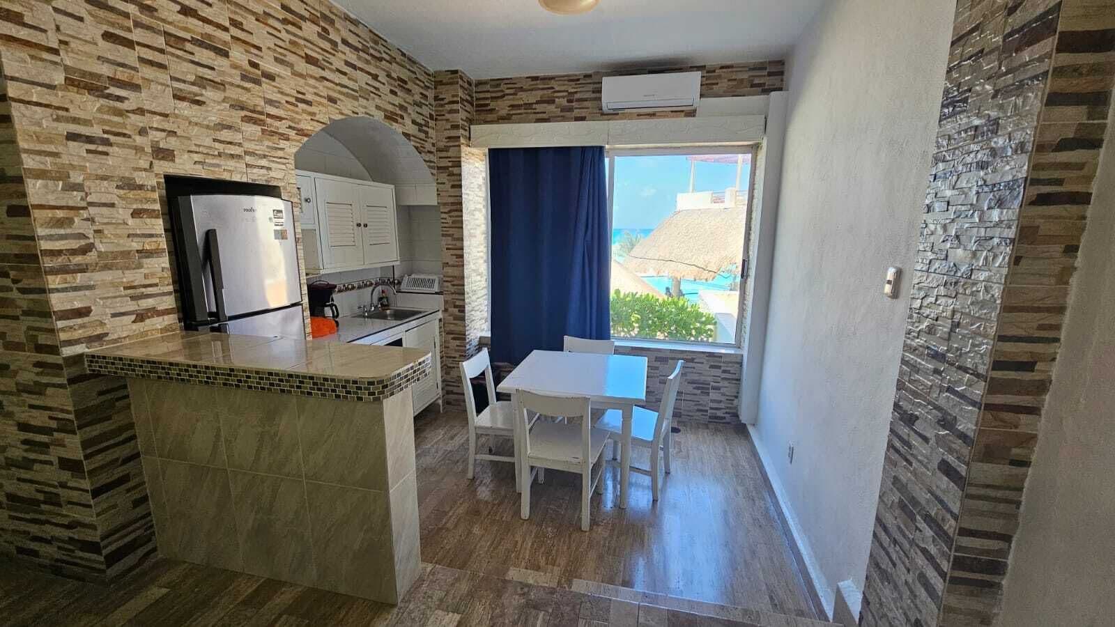 Eine kleine Küche mit integriertem Kühlschrank, Kaffeemaschine und Spüle. Es gibt eine kleine Essecke mit Tisch und Stühlen sowie ein Fenster mit Blick auf einen Pool und Gebäude.