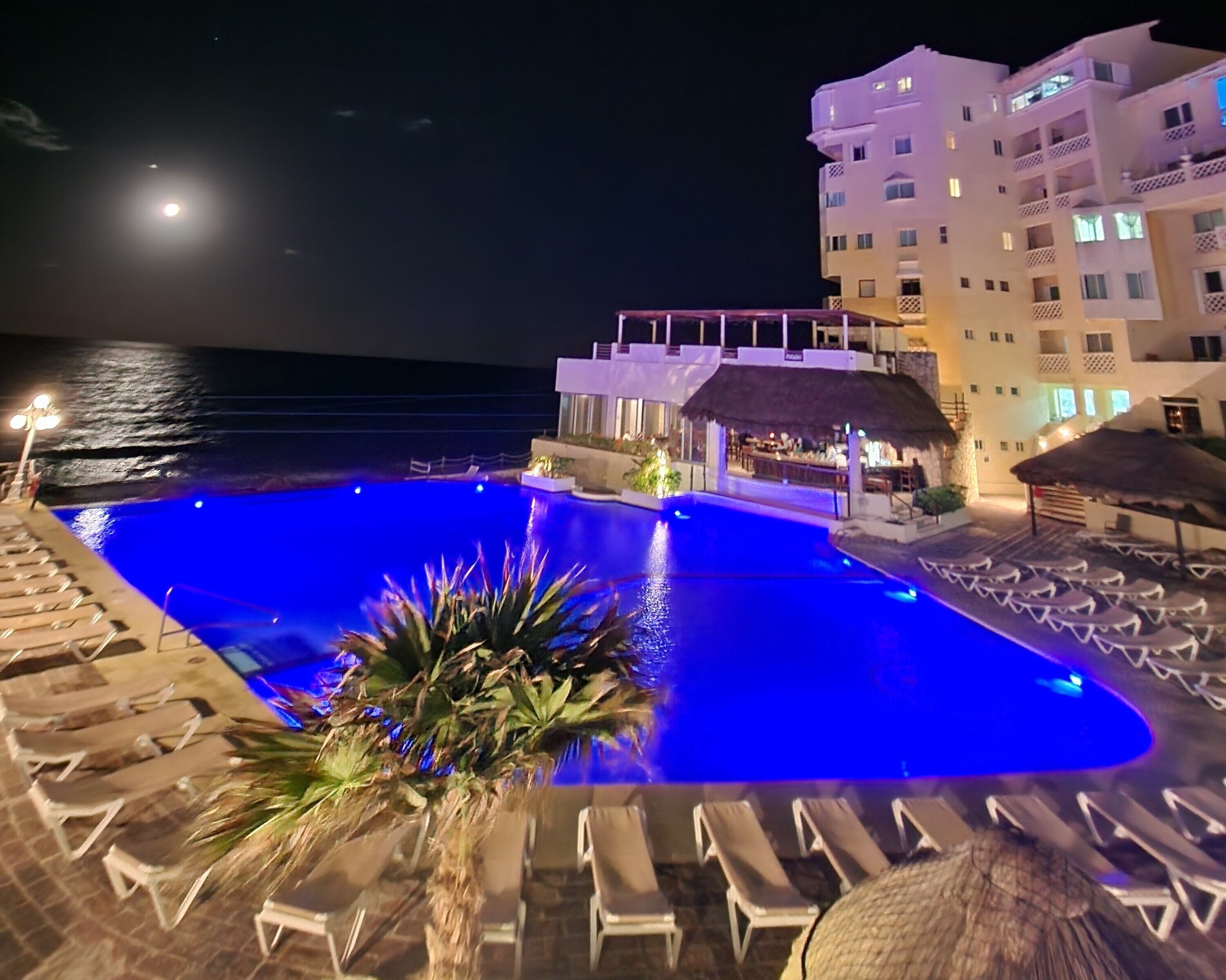 A nighttime pool area with lounge chairs and a bar, overlooking the sea.