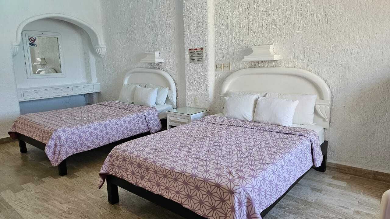 A hotel room with two beds, each with a white headboard and matching pillows. There is a small table between the beds, and a mirror above the headboard on the left.