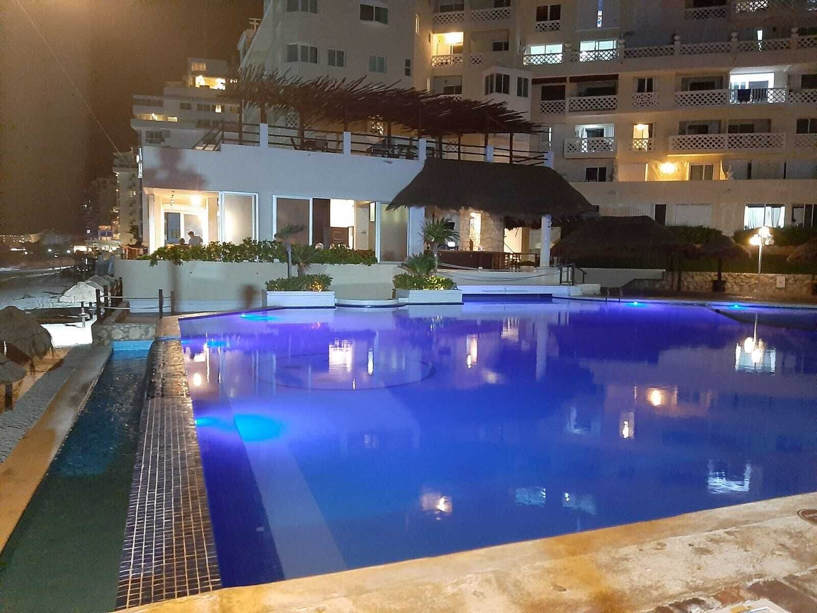 A nighttime view of a swimming pool with a tiled edge, surrounded by a building with balconies and a covered area with plants.