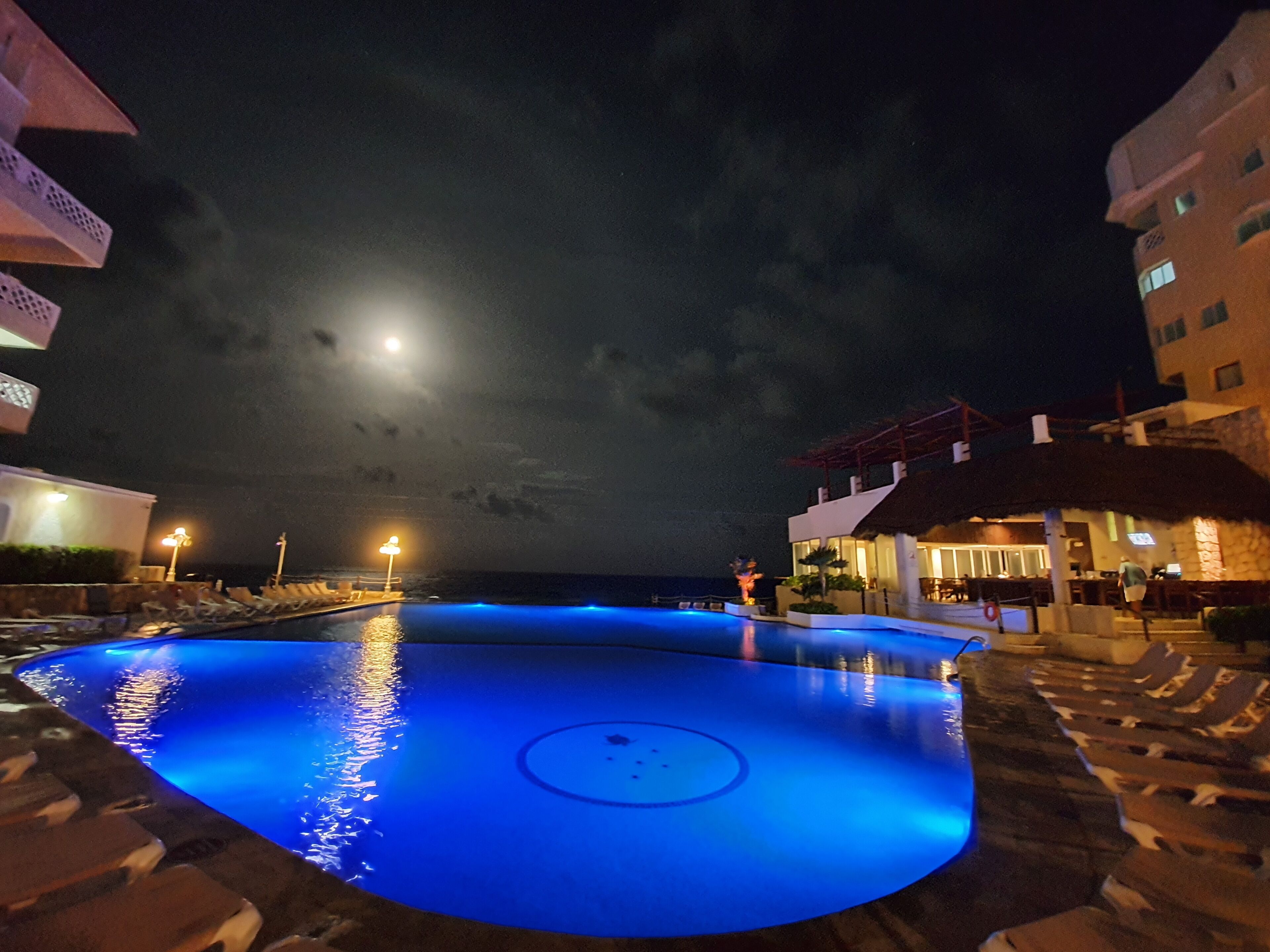 A nighttime pool area with a moonlit sky, a well-lit building, and a thatched-roof structure.