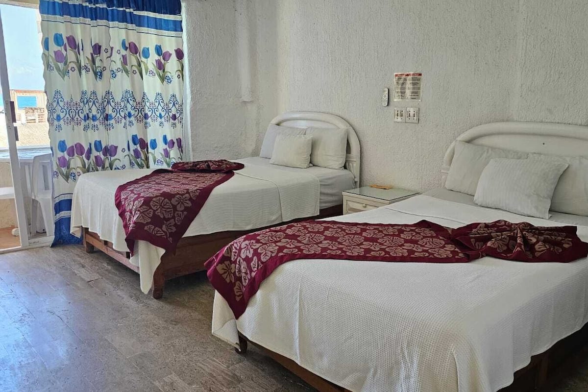 Deux lits simples avec du linge de lit blanc et des couvre-lits à motifs floraux rouges, dans une pièce dont un mur est d’un blanc uni et qui possède une fenêtre ornée d’un rideau à motifs floraux.