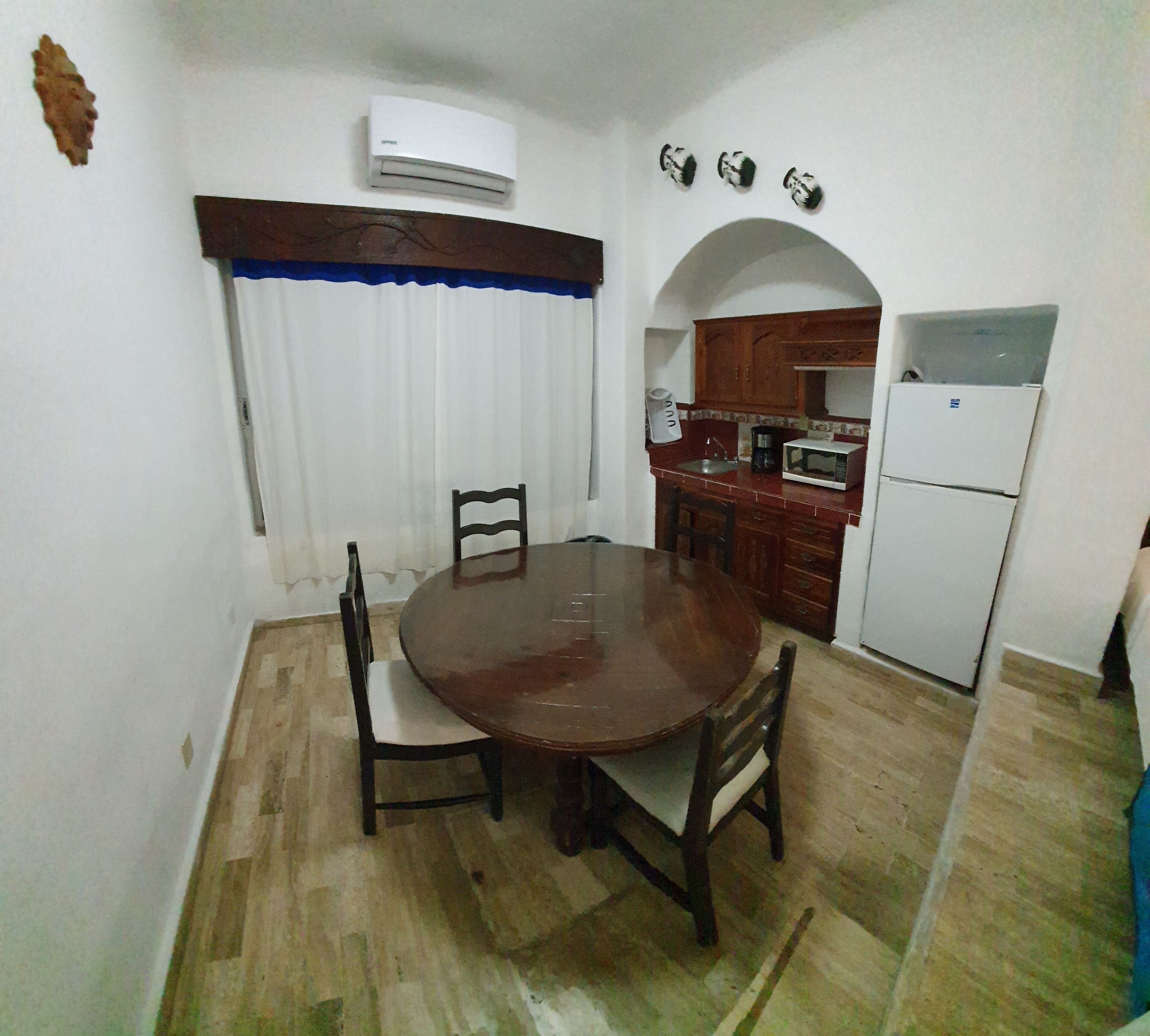 A compact kitchen with a dining area, featuring a round wooden table, chairs, a white refrigerator, a microwave, and a coffee maker.