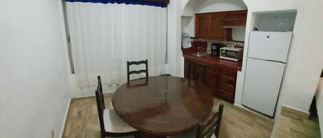 A compact kitchen with a dining area, featuring a round wooden table, chairs, a white refrigerator, a microwave, and a coffee maker.