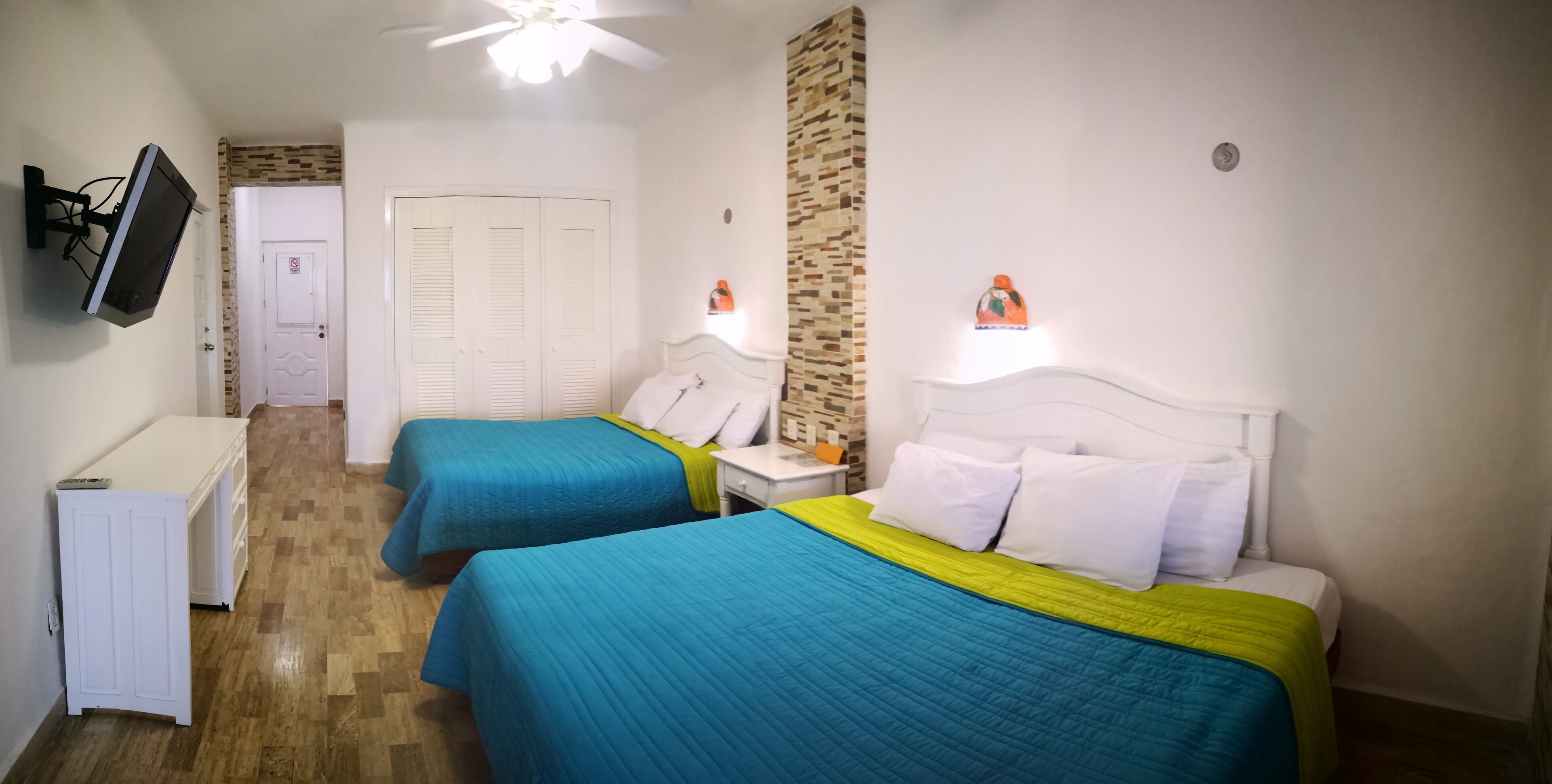 Une chambre d’hôtel avec deux lits, un téléviseur fixé au mur, un ventilateur de plafond et une petite table avec une lampe.