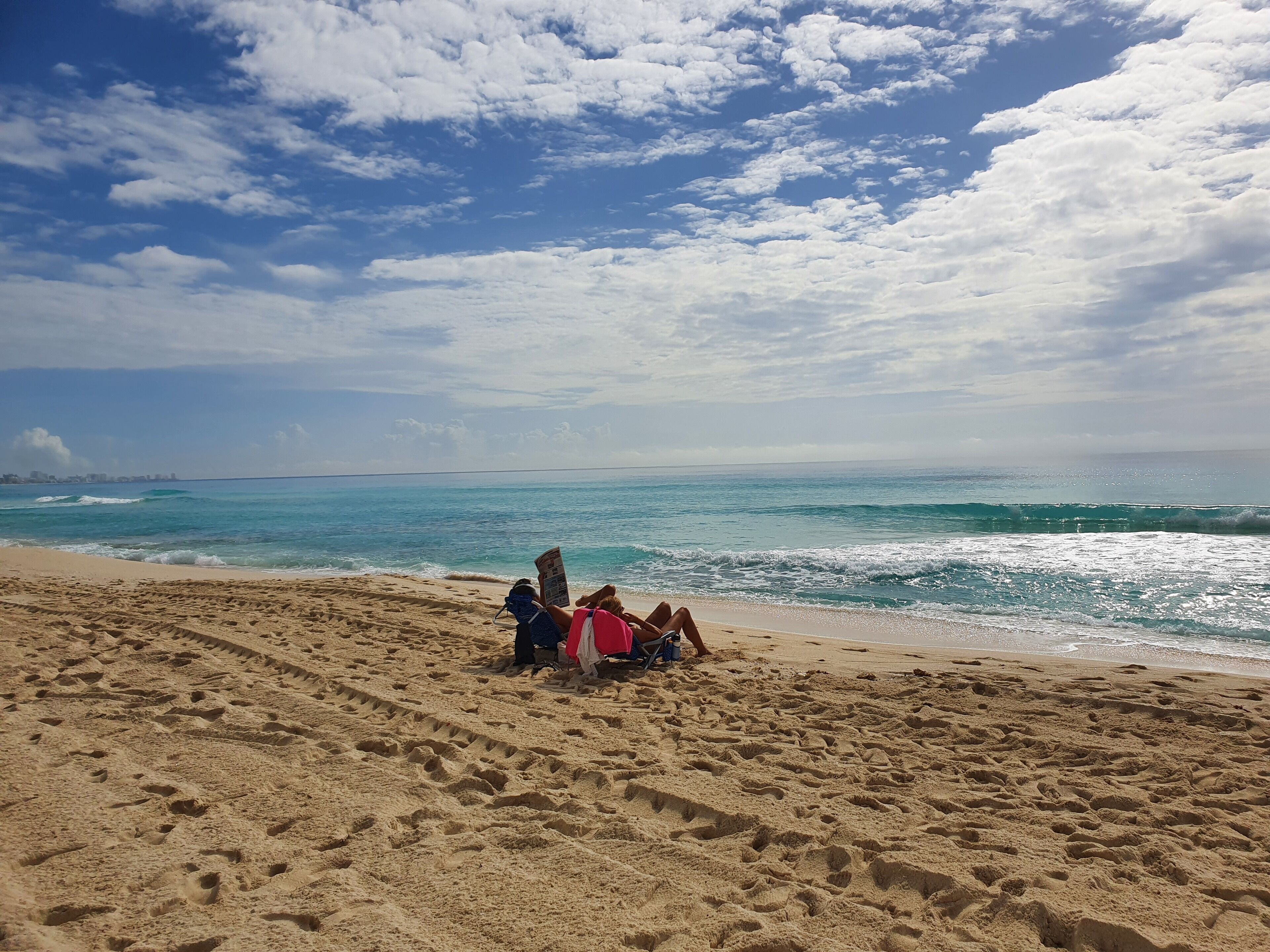 Una spiaggia sabbiosa con impronte che conducono all'acqua, cielo sereno e una linea di costa urbana in lontananza.